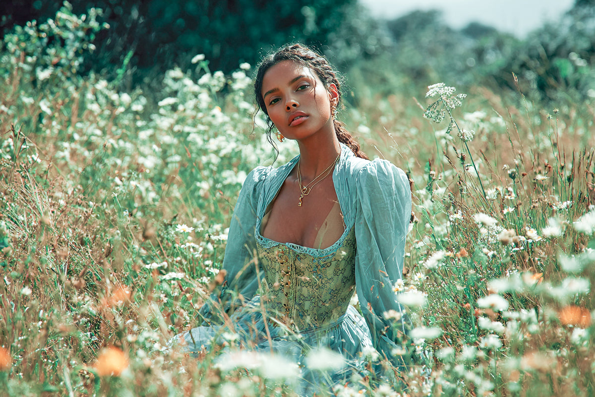 A person wearing a light blue vintage-style dress and layered necklaces sits among tall grasses and white wildflowers in a sunlit meadow, with trees softly blurred in the background.